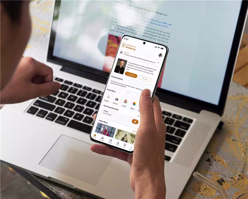 "User holding a smartphone with the Kristapp Church Management Software app open, showing the Archdiocese of Wellspring dashboard with announcements, quick-access icons, and Mass schedules, with a laptop in the background