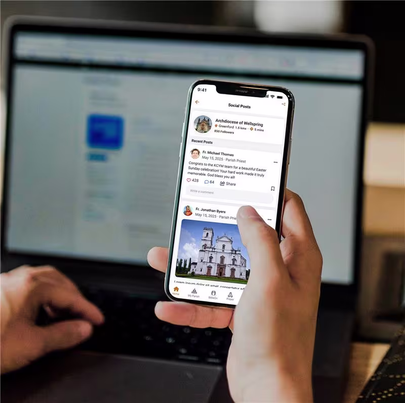 Person using a smartphone to browse parish social posts from the Archdiocese of Wellspring on the Kristapp Church Management Software mobile app, with a blurred laptop screen in the background.
